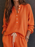 NEWYORKLISTED STYLE & COMFORT COLLECTION: A woman wearing a bright orange waffle-knit two-piece loungewear set, featuring a long-sleeve henley top with white buttons and matching drawstring pants, set against a dark textured wall.