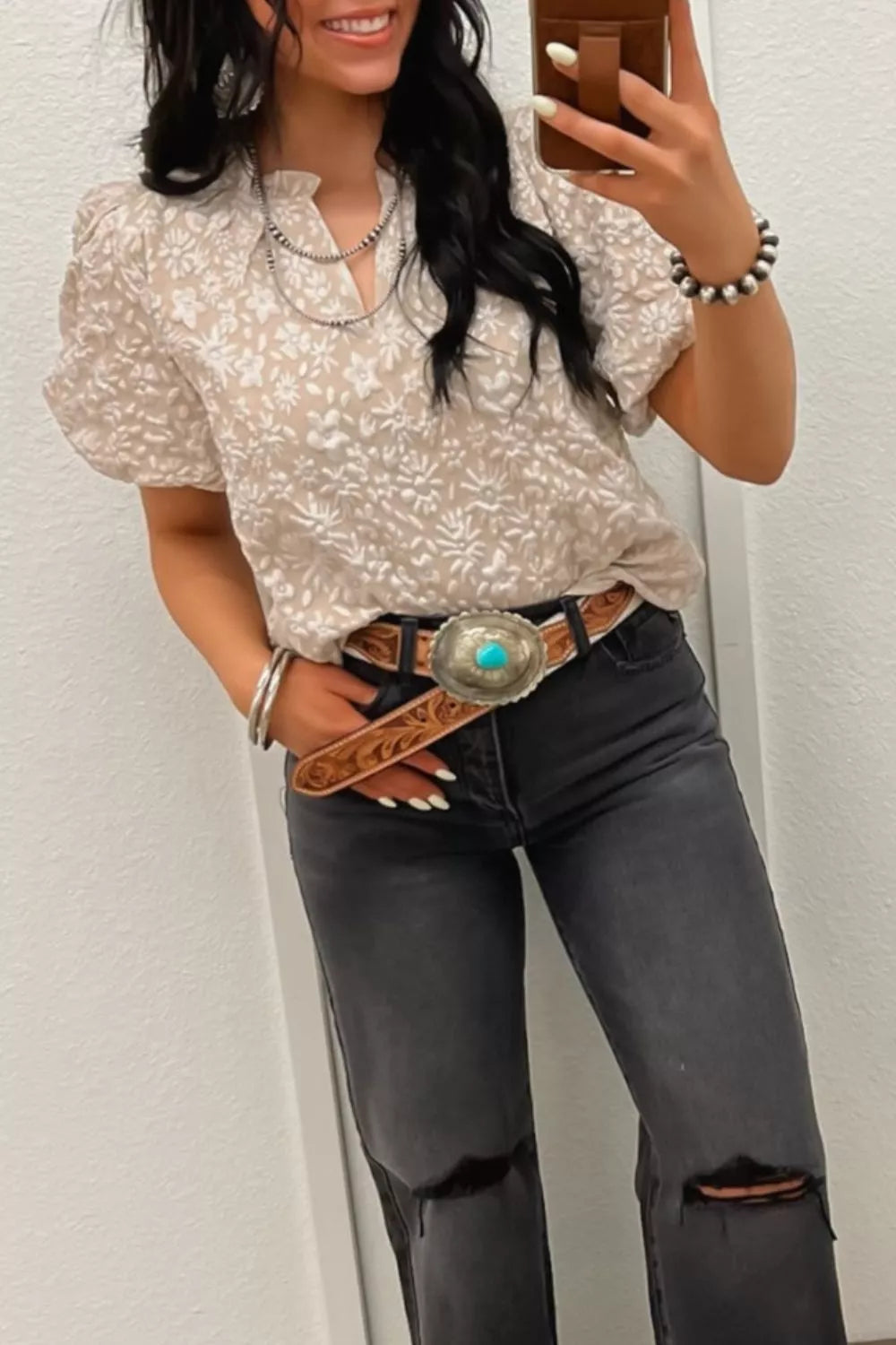 NEWYORKLISTED STYLE COLLECTION: Woman wearing a patterned blouse and black jeans with a belt, taking a mirror selfie.