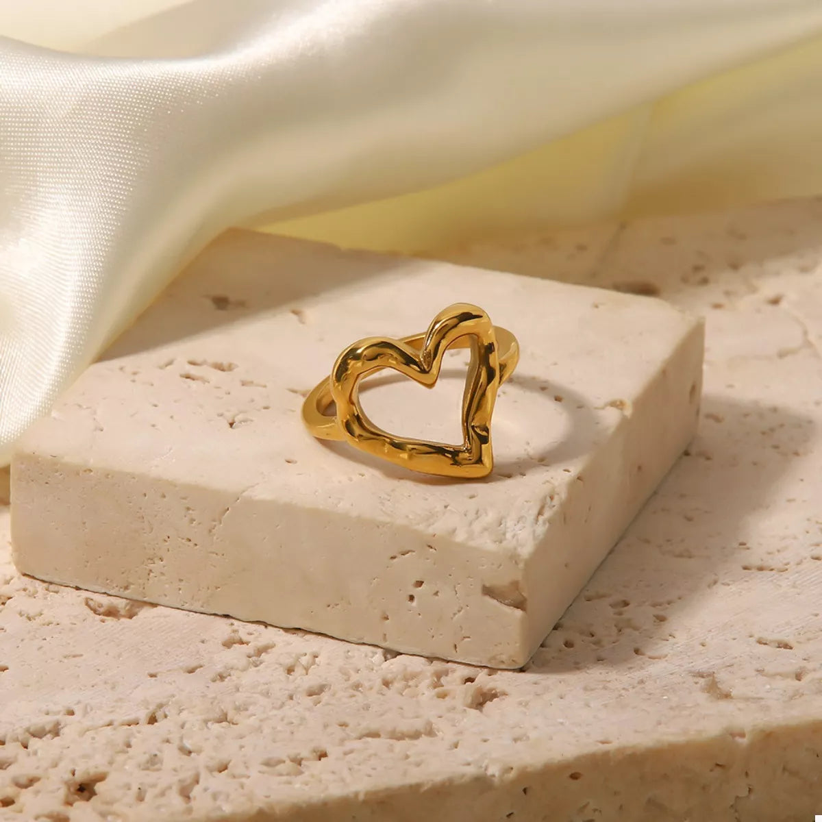 NYLD Glamour Collection by NEWYORKLISTED: Gold heart-shaped ring on a beige stone surface with a soft fabric background