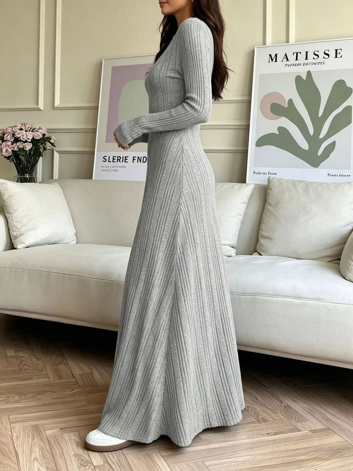 NEWYORKLISTED STYLE COLLECTION: A woman models a light gray ribbed knit maxi dress with long sleeves and a V-neckline in a bright, modern living room setting.