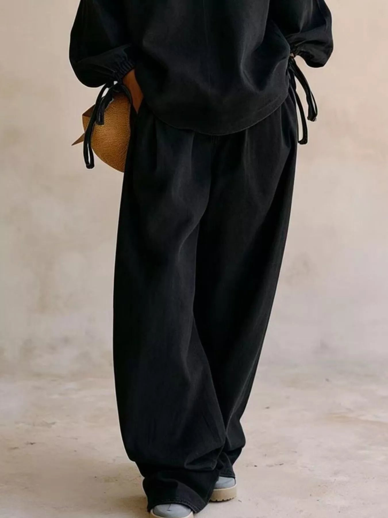 NEWYORKLISTED STYLE COLLECTION: A person wearing loose black drawstring pants and a matching black top, holding a small tan woven bag, photographed against a soft beige studio backdrop.