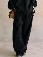 NEWYORKLISTED STYLE COLLECTION: A person wearing loose black drawstring pants and a matching black top, holding a small tan woven bag, photographed against a soft beige studio backdrop.