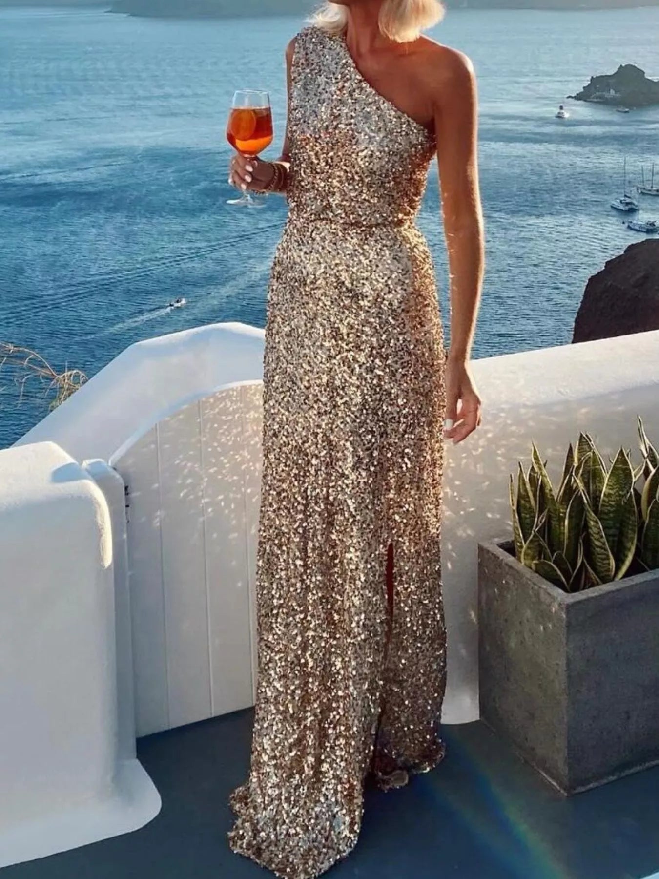 NEWYORKLISTED STYLE COLLECTION: Woman in a sparkly one-shoulder dress holding a glass of wine by the ocean.