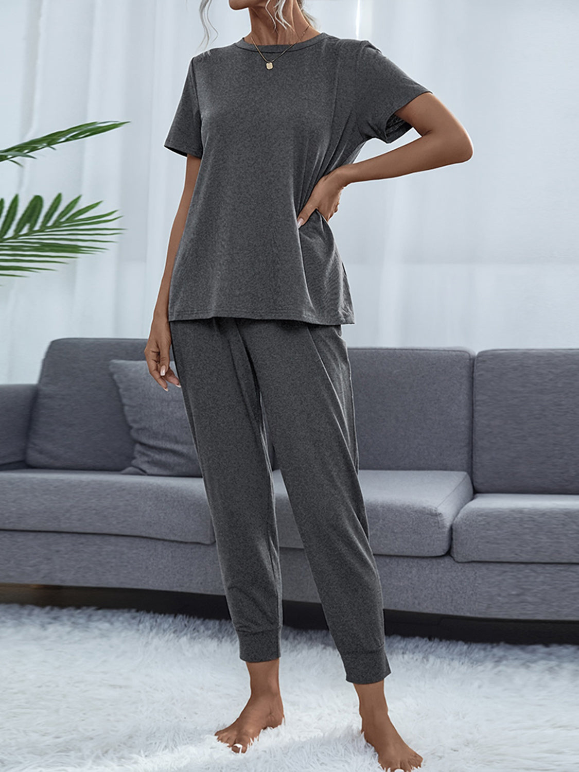 NEWYORKLISTED Intimate Apparel: Person wearing a gray pajama set standing in a living room with a gray sofa and white curtains.