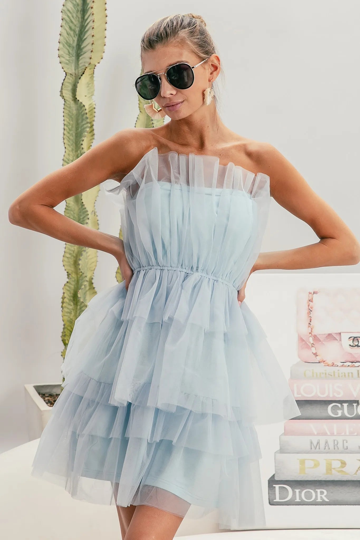 NEWYORKLISTED STYLE COLLECTION: Sleeveless Layered Organza Dress Woman wearing a light blue dress and sunglasses, standing in front of a stack of books with visible brand names.