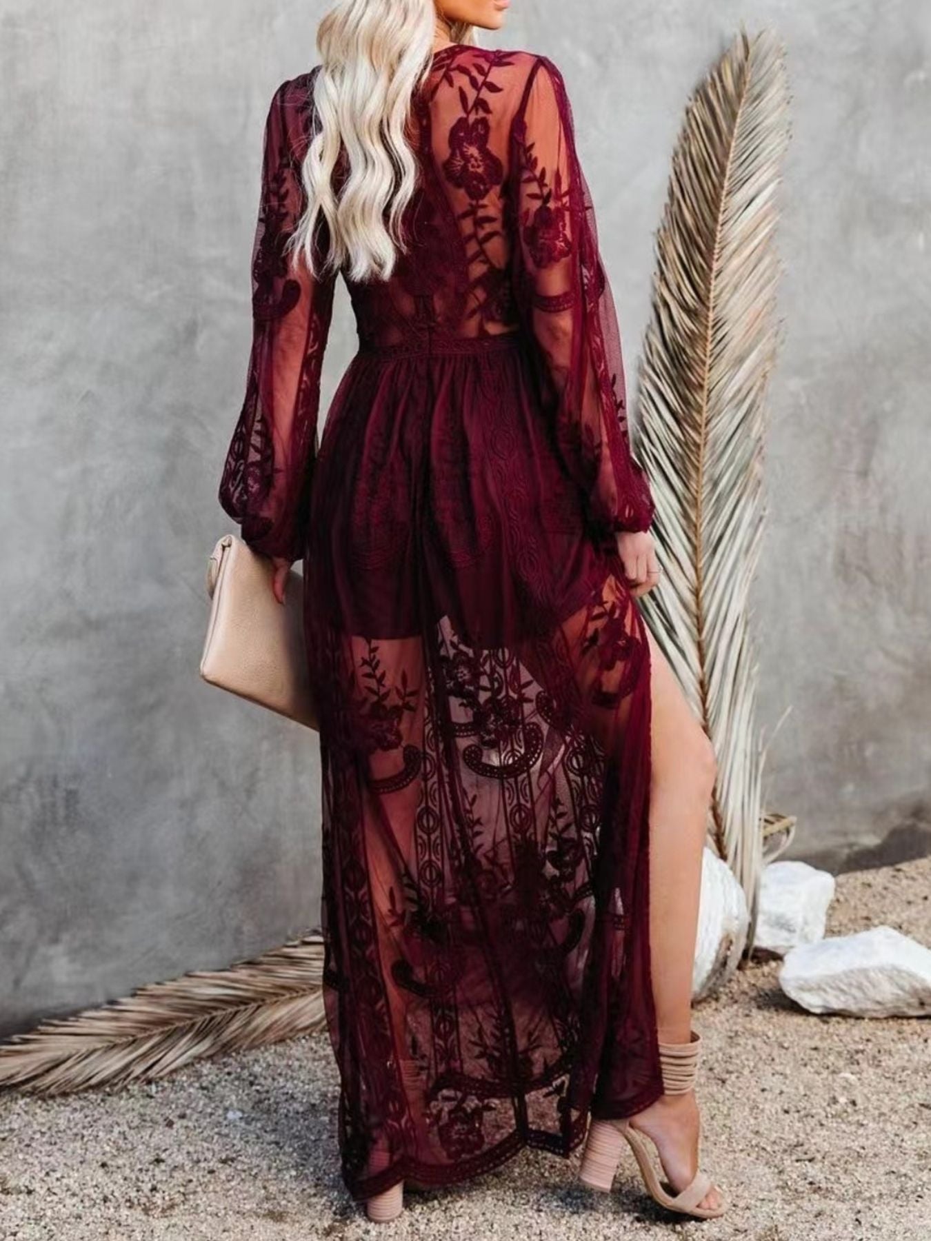 NEWYORKLISTED STYLE COLLECTION: Woman wearing a burgundy lace dress with long sleeves and a slit, standing against a textured wall.