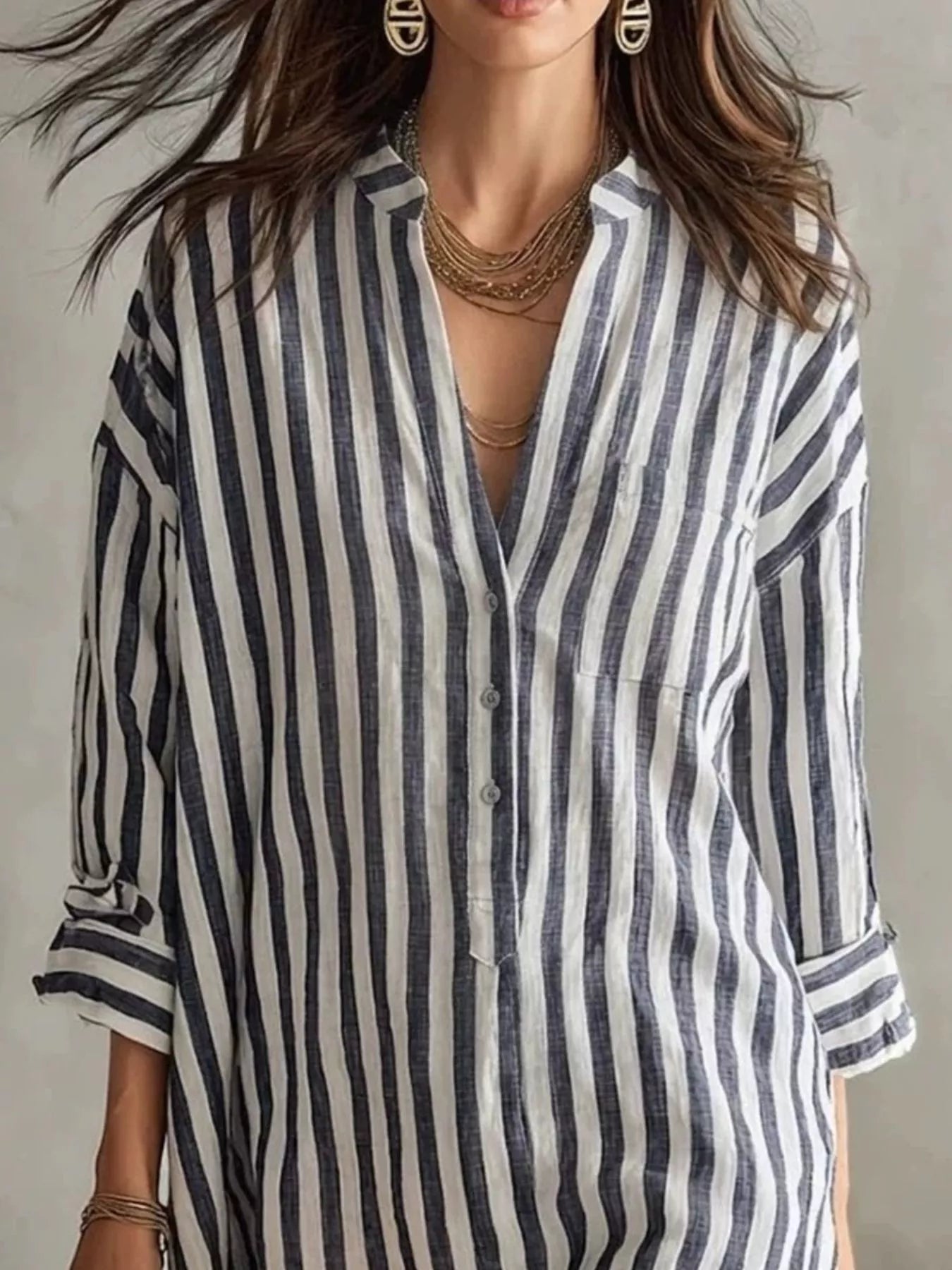 NEWYORKLISTED STYLE COLLECTION: A woman wearing a loose-fitting linen shirt dress with vertical navy and off-white stripes, styled with layered gold necklaces and earrings.