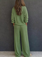 NEWYORKLISTED STYLE & COMFORT COLLECTION: A woman viewed from behind wearing a matching sage green knit lounge set consisting of a long-sleeve top and wide-leg pants, standing against a dark gray textured wall.