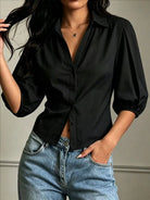 NEWYORKLISTED STYLE COLLECTION: A woman wearing a black button-up blouse with three-quarter puff sleeves and light-wash blue jeans, standing against a neutral gray wall.