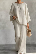 NEWYORKLISTED STYLE COLLECTION: A woman wearing a loose, two-piece linen outfit in a warm off-white or cream color, consisting of a wide-sleeve top and matching wide-leg pants. She is accessorized with layered silver necklaces, a silver watch, and holds a small woven straw handbag against a textured gray studio background.