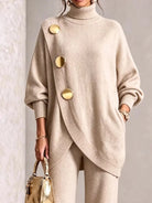 NEWYORKLISTED STYLE COLLECTION:  A woman wearing a beige oversized turtleneck sweater with large gold-tone decorative buttons and matching beige pants, holding a small gold handbag against a light textured wall.