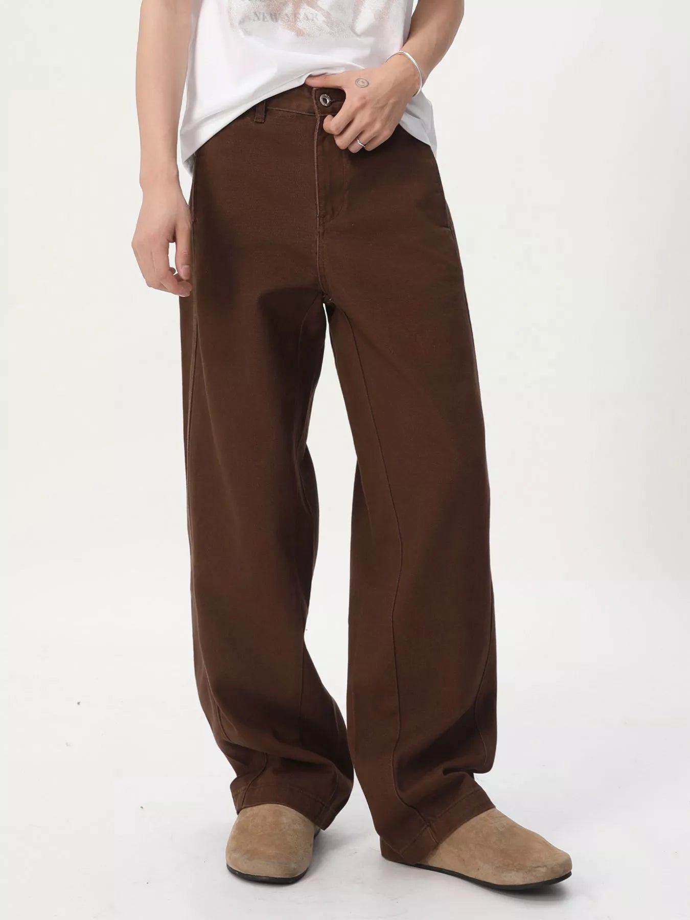 NEWYORKLISTED ATLAS MEN'S COLLECTION: A person wearing loose brown wide-leg pants with a white graphic t-shirt and beige suede shoes, standing against a plain white studio background.