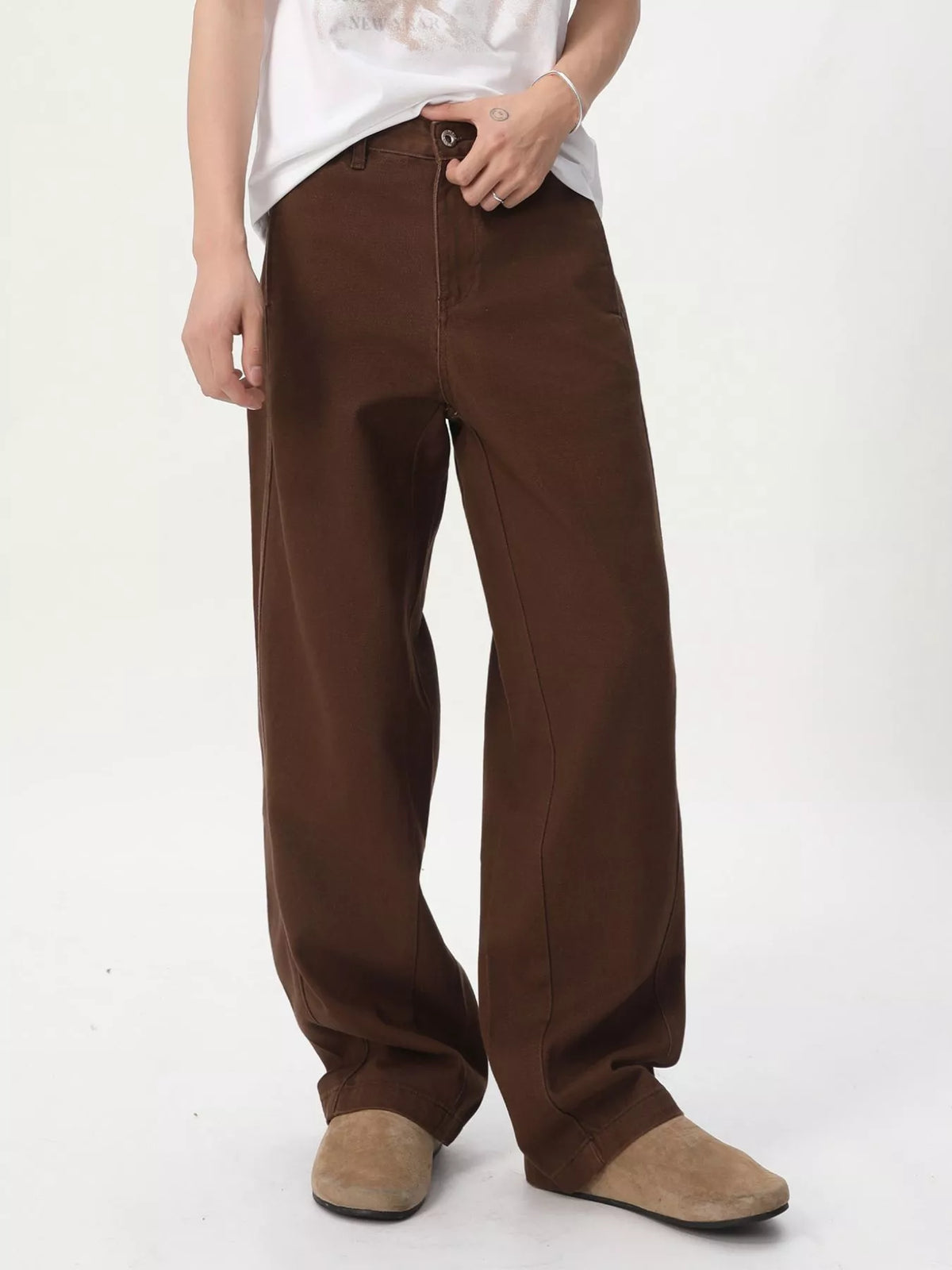 NEWYORKLISTED ATLAS MEN'S COLLECTION: A person wearing loose brown wide-leg pants with a white graphic t-shirt and beige suede shoes, standing against a plain white studio background.