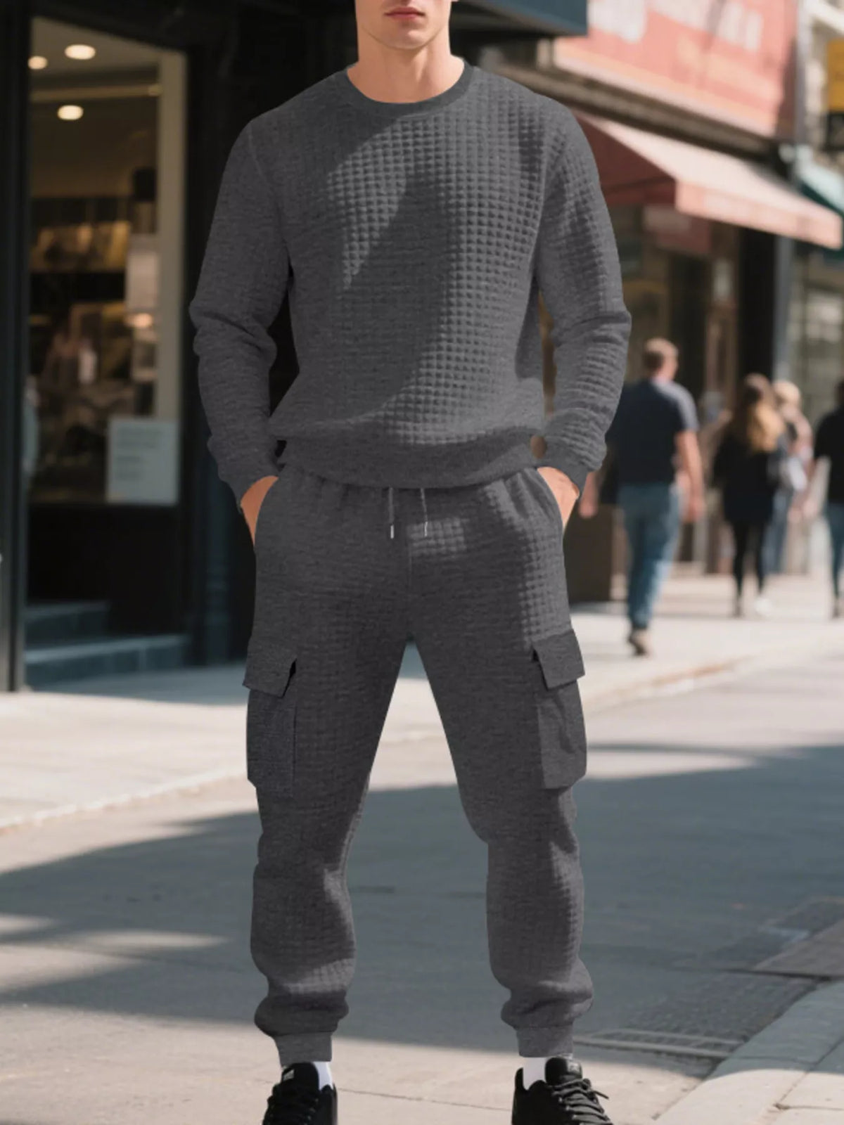 NEWYORKLISTED ATLAS MEN'S COLLECTION: A man wearing a matching dark gray waffle-knit crewneck sweatshirt and jogger cargo pants stands on a city sidewalk with blurred storefronts and pedestrians in the background.