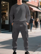 NEWYORKLISTED ATLAS MEN'S COLLECTION: A man wearing a matching dark gray waffle-knit crewneck sweatshirt and jogger cargo pants stands on a city sidewalk with blurred storefronts and pedestrians in the background.