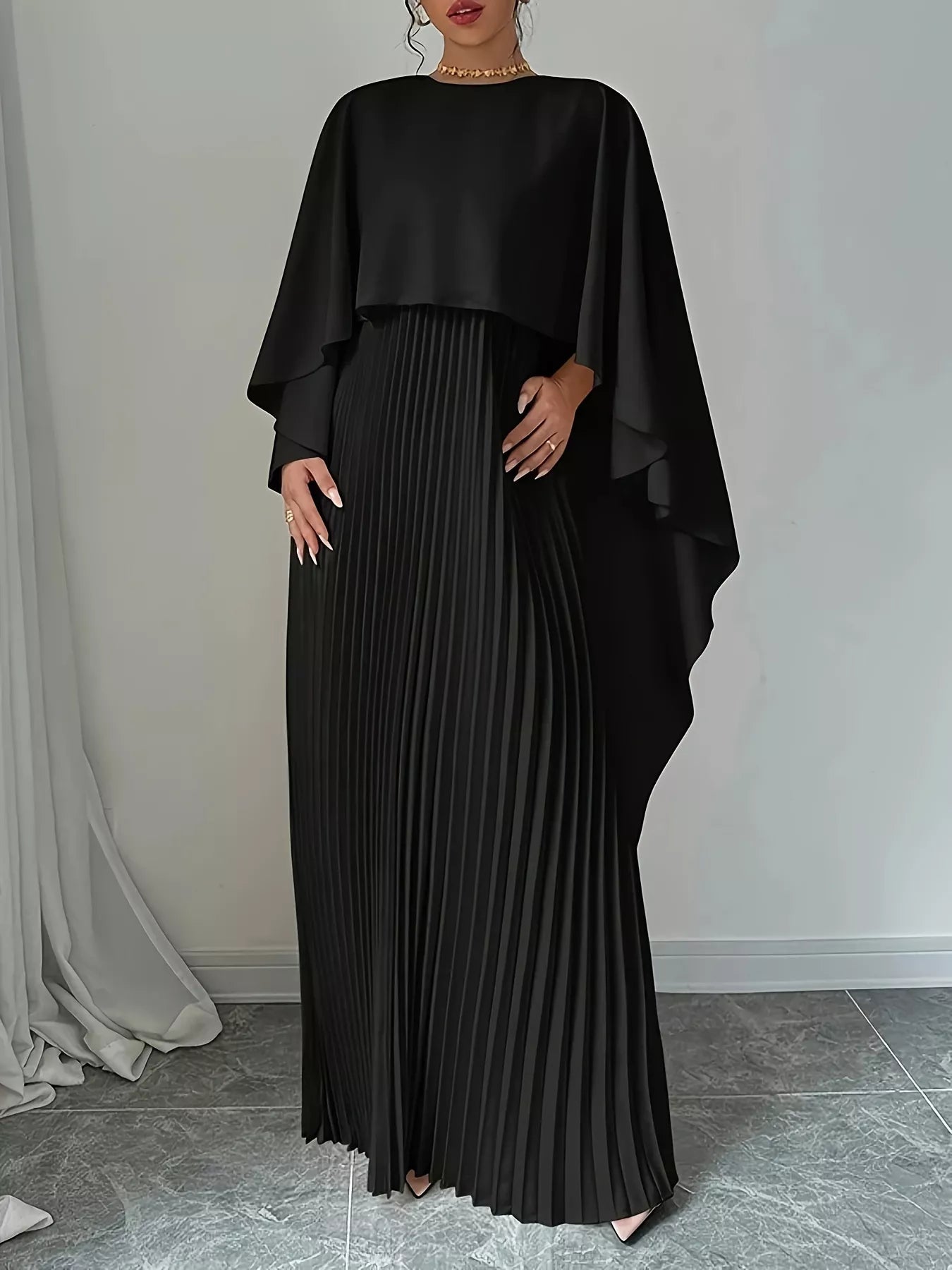 NEWYORKLISTED STYLE COLLECTION: Woman wearing a black pleated dress with a cape-like top against a neutral background