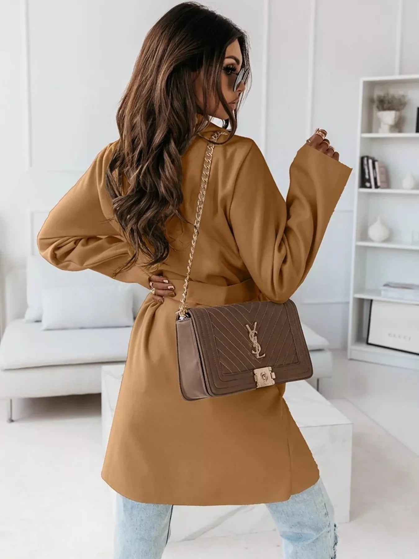 Woman in a tan coat holding a brown handbag in a modern living room.