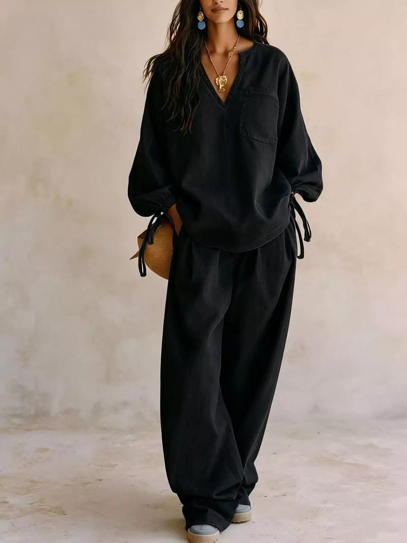 NEWYORKLISTED STYLE COLLECTION: A woman models a black two-piece lounge set consisting of a long-sleeve V-neck top with tie details at the cuffs and matching wide-leg pants, set against a textured beige wall.