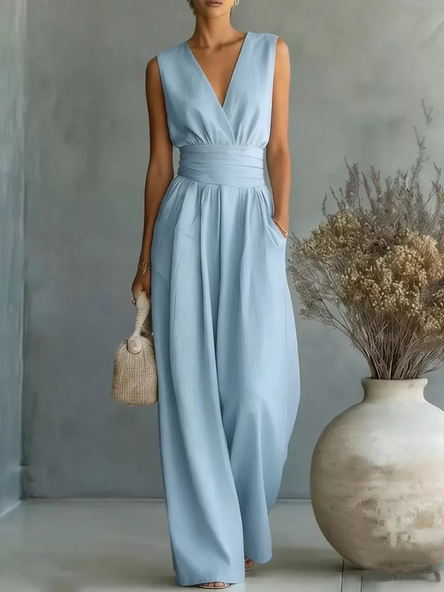 NEWYORKLISTED STYLE COLLECTION: Woman wearing a light blue dress holding a woven bag in a minimalistic setting with a vase and dried plants.