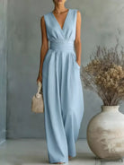 NEWYORKLISTED STYLE COLLECTION: Woman wearing a light blue dress holding a woven bag in a minimalistic setting with a vase and dried plants.