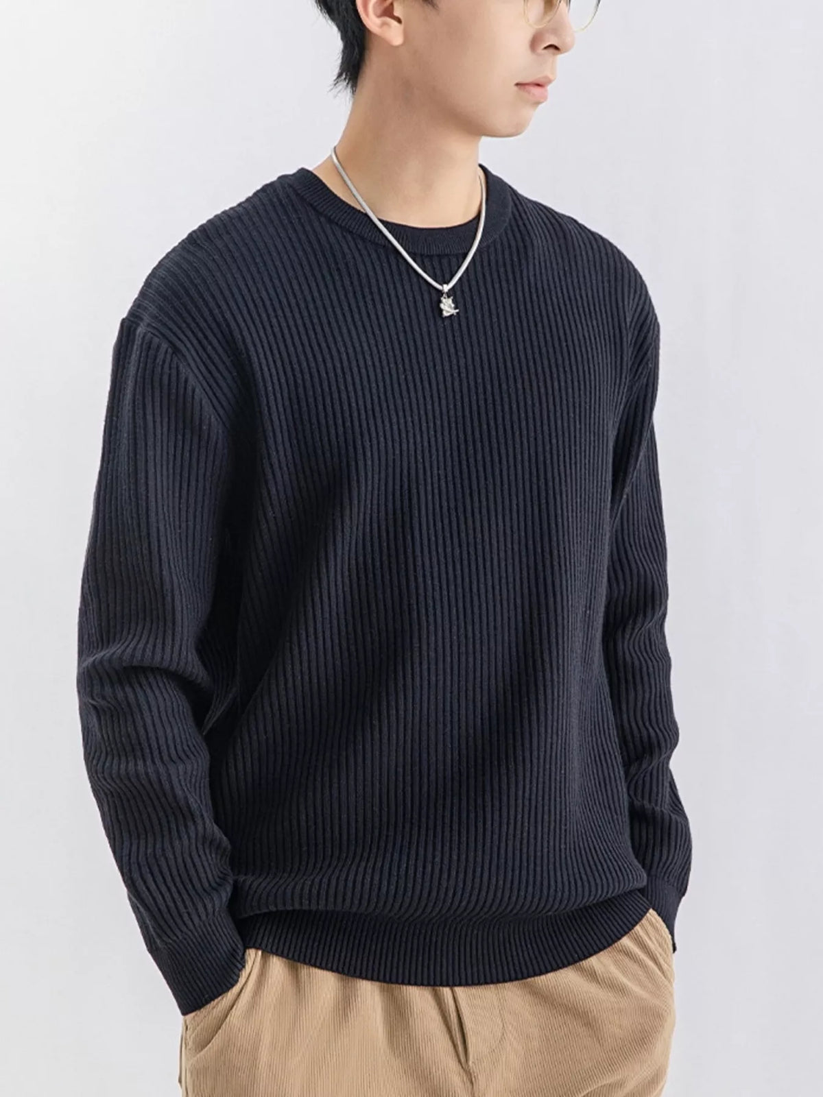 NEWYORKLISTED ATLAS MEN'S COLLECTION: A man wearing a dark navy ribbed knit crewneck sweater, paired with beige pants and a silver necklace, photographed against a light gray studio background.