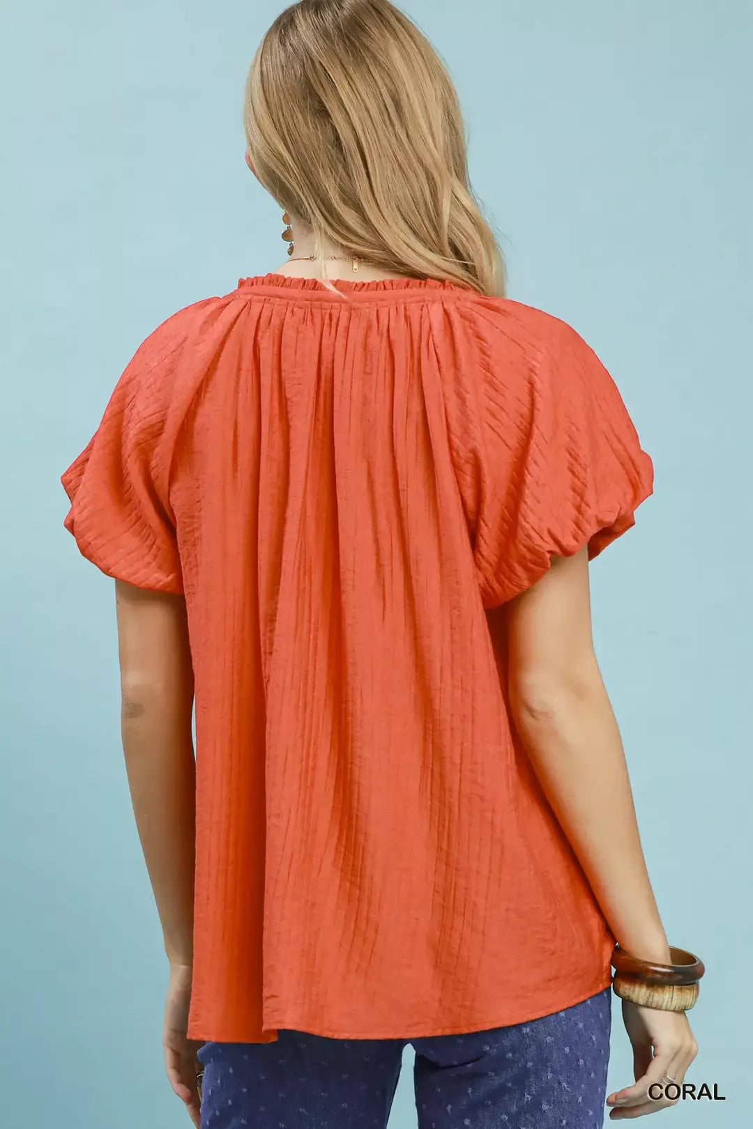 NEWYORKLISTED STYLE COLLECTION: Woman wearing a coral blouse with short sleeves against a light blue background