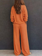 NEWYORKLISTED STYLE & COMFORT COLLECTION: A woman viewed from behind wearing a matching burnt orange knit lounge set consisting of a long-sleeve top and wide-leg pants, standing against a dark gray textured wall.