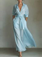 NEWYORKLISTED STYLE COLLECTION: A woman models a light blue wrap-style maxi dress with a tie waist, long sleeves rolled to the forearm, and side pockets, photographed against a neutral gray studio backdrop.