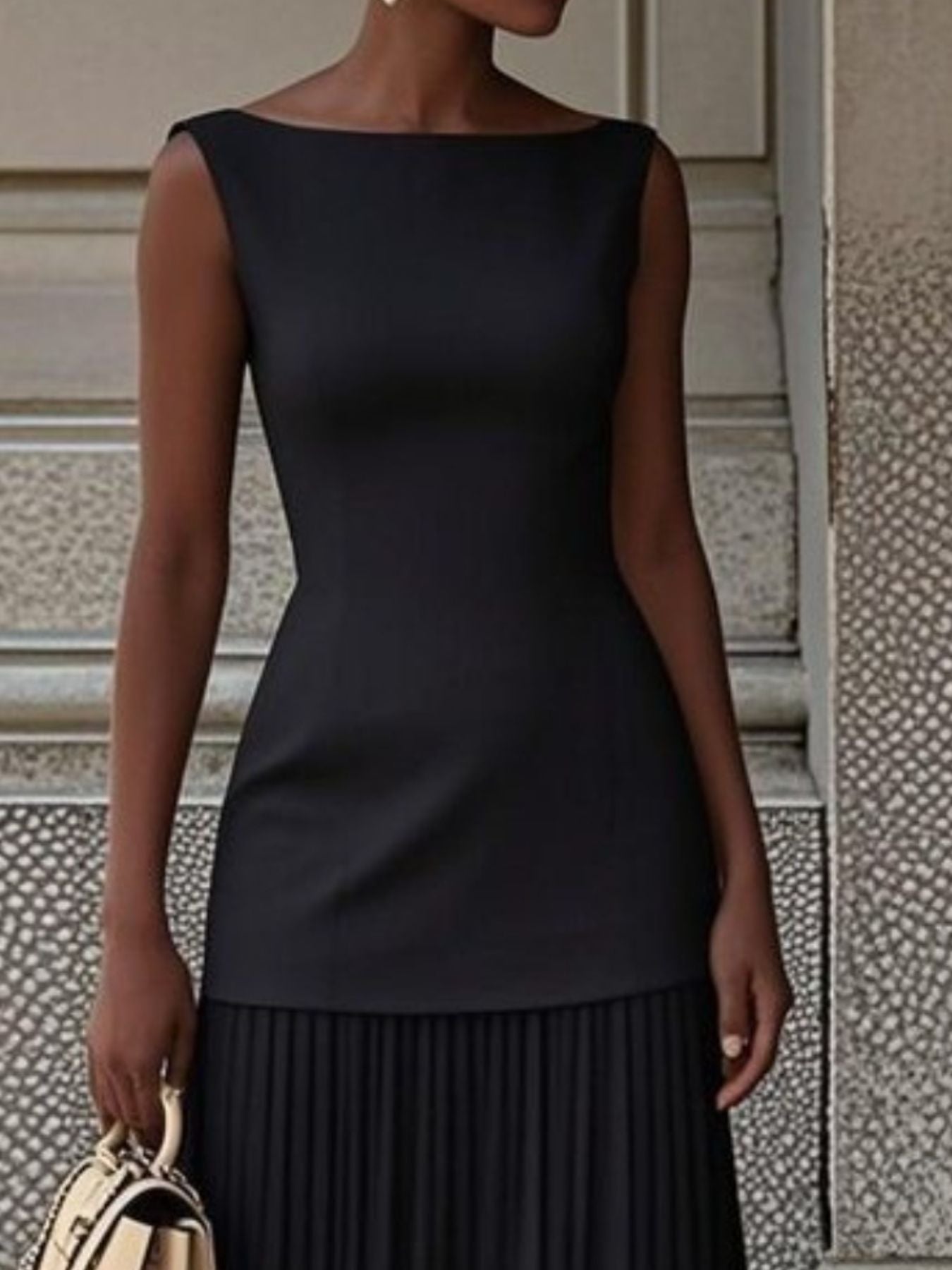 NEWYORKLISTED STYLE COLLECTION: Woman wearing a black dress with pleated skirt against a textured wall.