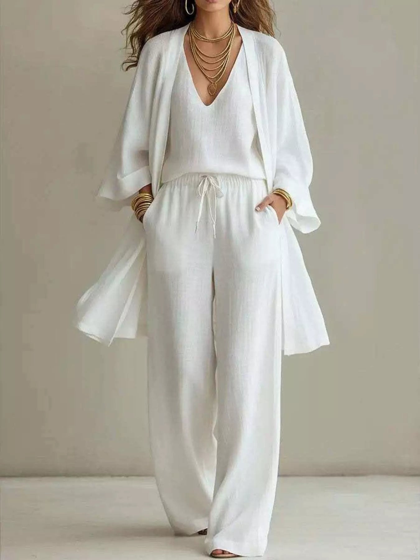 NEWYORKLISTED STYLE COLLECTION: A woman modeling a white three-piece linen outfit consisting of a V-neck top, matching wide-leg pants with a drawstring, and a lightweight open-front cardigan. She is accessorized with layered gold necklaces and bracelets, standing against a neutral beige studio background.