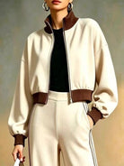 NEWYORKLISTED STYLE COLLECTION: A woman models a cream-colored bomber jacket with dark brown ribbed trim, paired with matching cream pants and a black top, set against a neutral studio background.