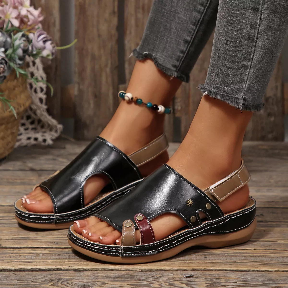 NEWYORKLISTED SHOE COLLECTION: A pair of women's black leather platform slide sandals with cutout toes, beige ankle straps, and decorative gold-tone studs, shown on a model's feet on a rustic wooden floor.