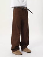 NEWYORKLISTED ATLAS MEN'S COLLECTION: A person shown from the waist down wearing loose brown wide-leg pants, a black belt, a white shirt, and tan slip-on shoes against a plain light background.
