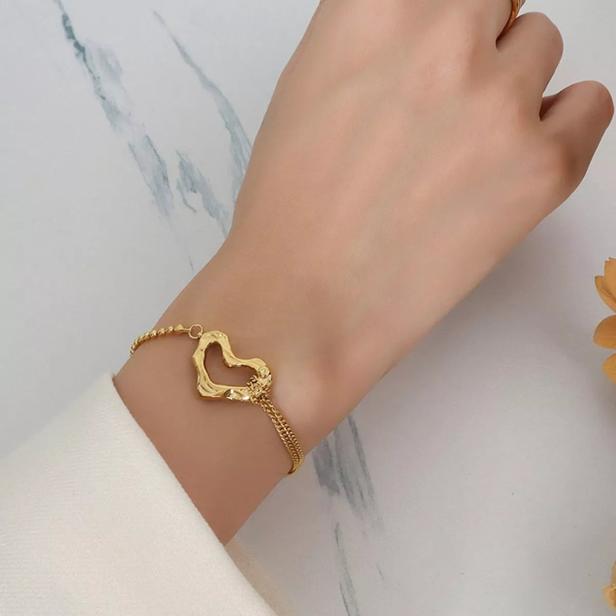 NYLD Glamour Collection by NEWYORKLISTED: Gold bracelet with a heart design on a wrist against a marble background