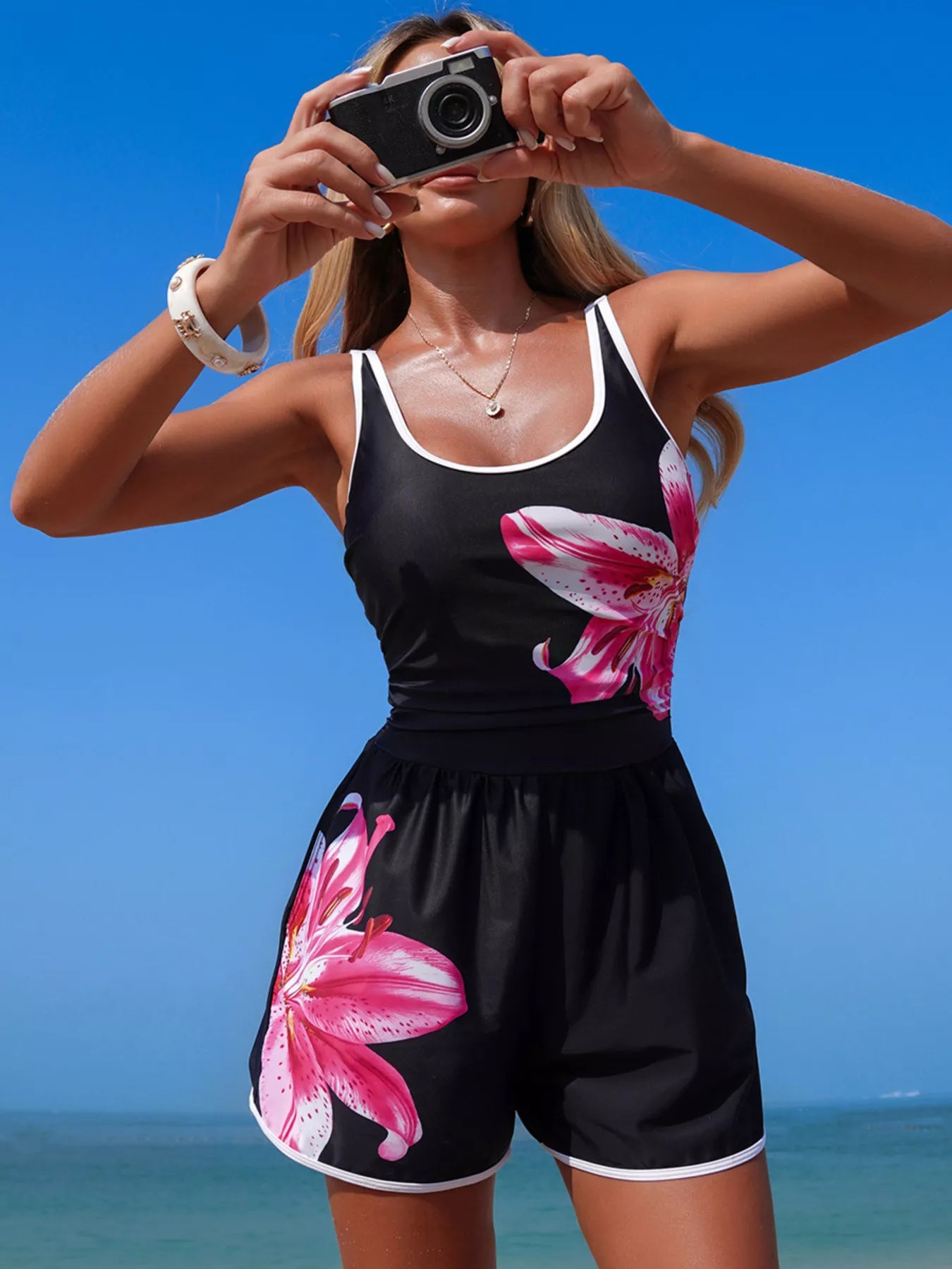 NEWYORKLISTED STYLE COLLECTION: Woman taking a selfie on a beach wearing a black tank top and shorts with pink floral designs.