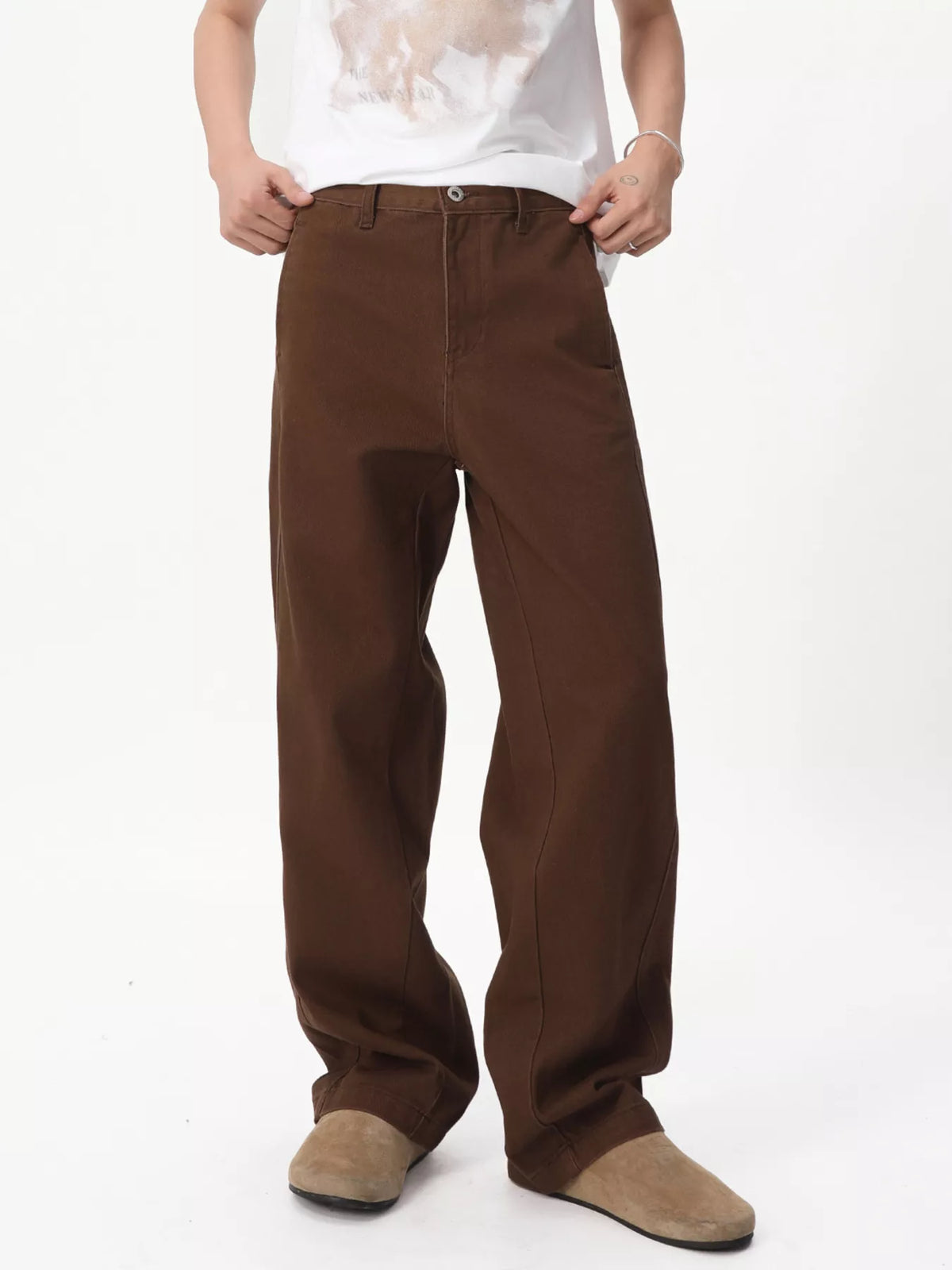 NEWYORKLISTED ATLAS MEN'S COLLECTION: A person wearing loose-fit brown wide-leg pants, styled with a white graphic t-shirt and beige slip-on shoes, photographed against a plain white studio background.