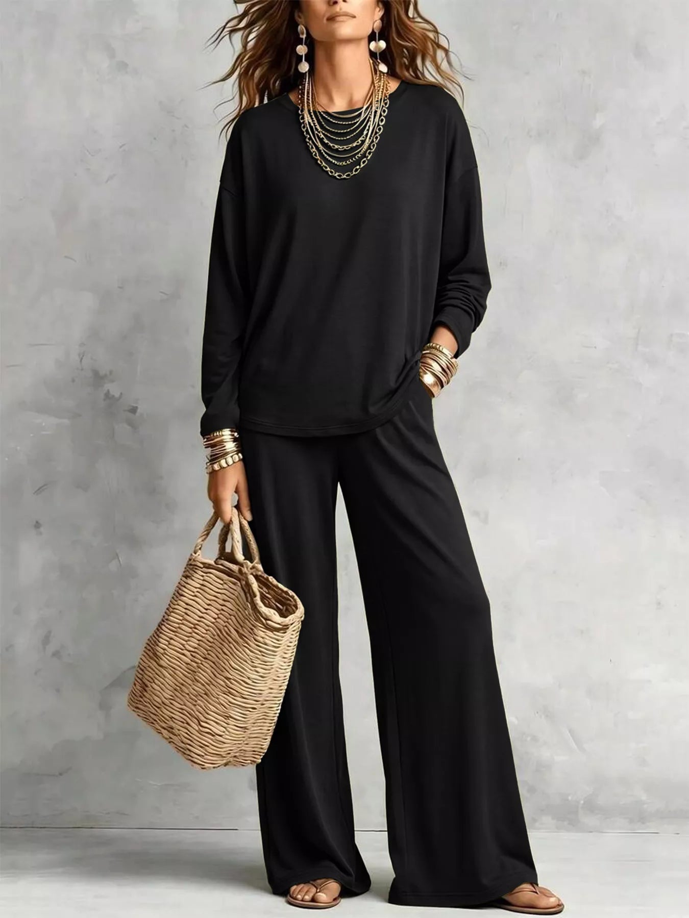 NEWYORKLISTED STYLE COLLECTION: A woman models a black long-sleeve top and matching wide-leg pants, styled with layered gold necklaces, bracelets, and a woven straw tote bag against a textured gray wall.