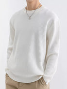 NEWYORKLISTED ATLAS MEN'S COLLECTION: A person wearing a ribbed long-sleeve crewneck sweater in off-white, paired with beige pants and a silver pendant necklace, against a plain light background.