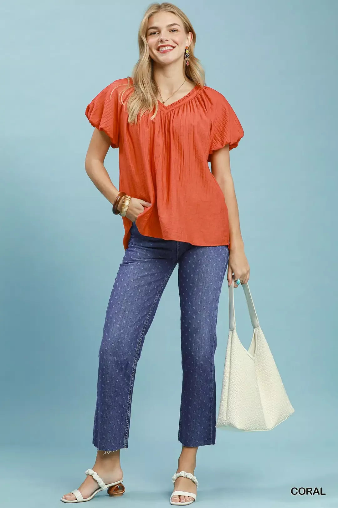 NEWYORKLISTED STYLE COLLECTION: Woman wearing an orange blouse and blue jeans holding a white bag on a light blue background