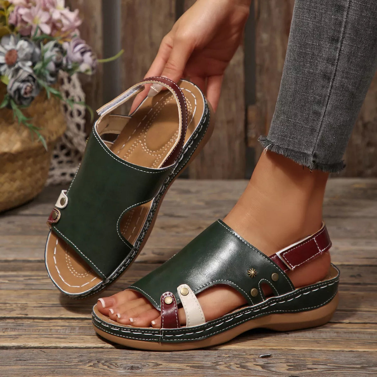 NEWYORKLISTED SHOE COLLECTION: A person wearing a pair of dark green leather slide sandals with burgundy straps and white contrast stitching, standing on a rustic wooden floor while holding the other sandal.