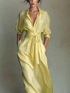 NEWYORKLISTED STYLE COLLECTION: A woman wearing a pale yellow wrap-style midi dress with three-quarter sleeves, a tie waist, and side pockets, photographed against a plain light gray studio background.
