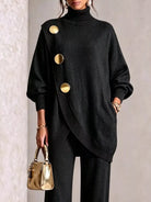 NEWYORKLISTED STYLE COLLECTION:  A woman models a black oversized turtleneck sweater with asymmetrical gold buttons, paired with black pants and holding a small metallic gold handbag.