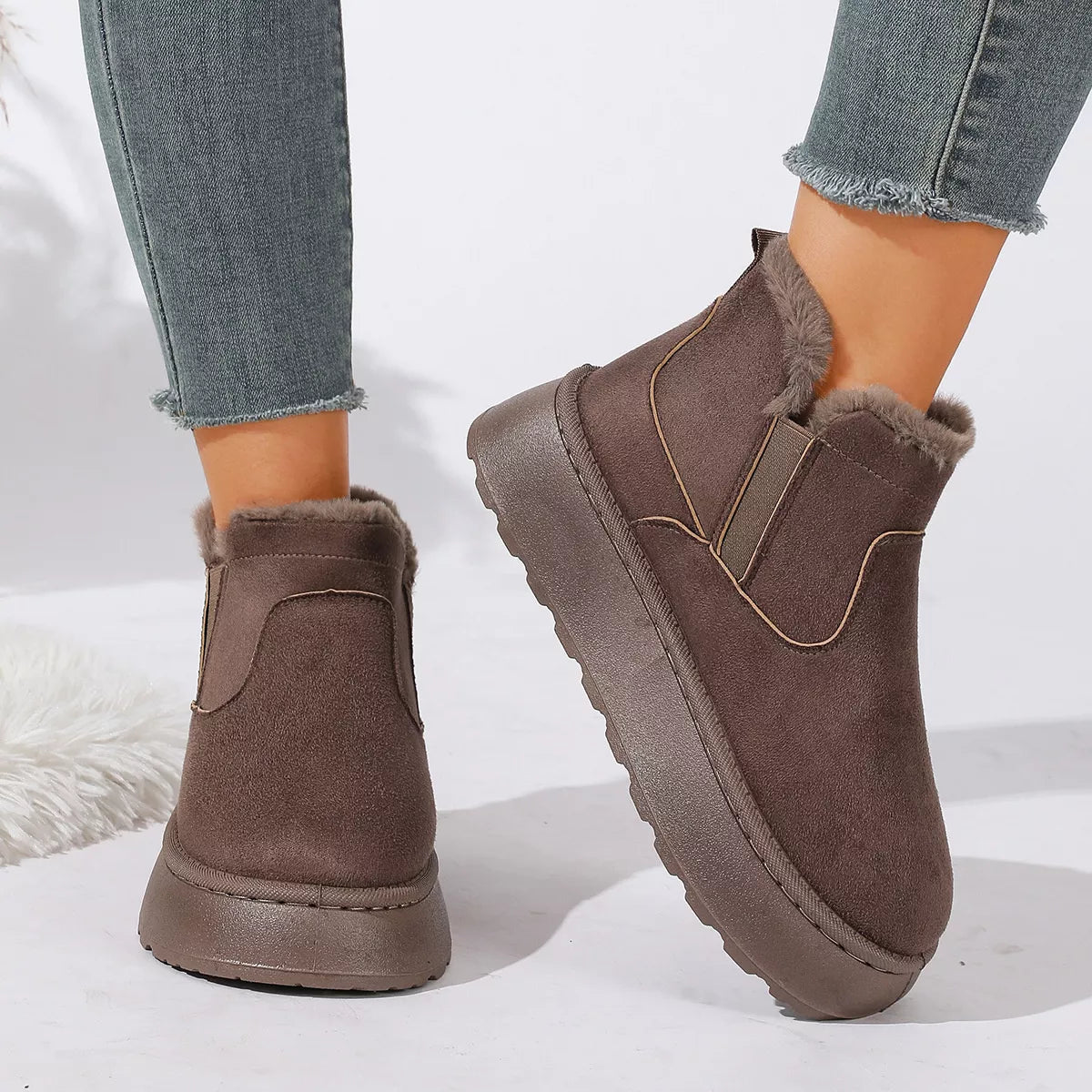 NEWYORKLISTED SHOE COLLECTION: A pair of women's brown suede-like ankle boots with plush faux-fur lining and elastic side panels, shown worn with gray frayed-hem jeans on a white studio background.