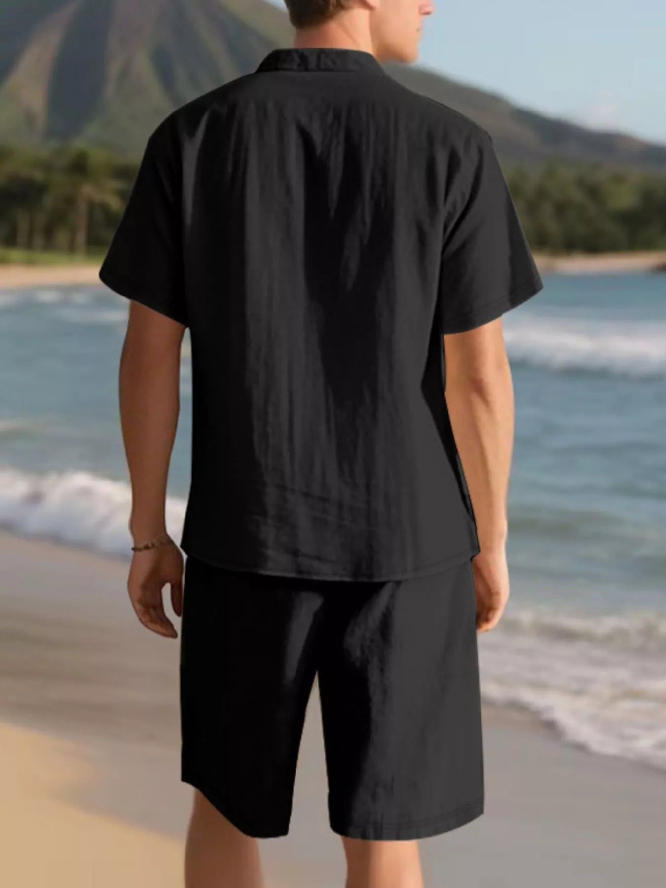 NEWYORKLISTED ATLAS MEN'S COLLECTION: Person wearing a black shirt and shorts standing on a beach with mountains in the background