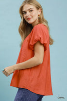 NEWYORKLISTED STYLE COLLECTION: Woman wearing a coral top with ruffled sleeves against a light blue background