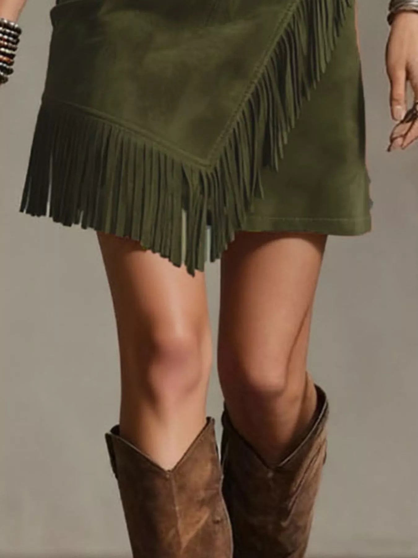 NEWYORKLISTED STYLE COLLECTION: An olive green faux-suede mini skirt with asymmetrical fringe trim, styled with brown suede cowboy boots against a neutral studio background.