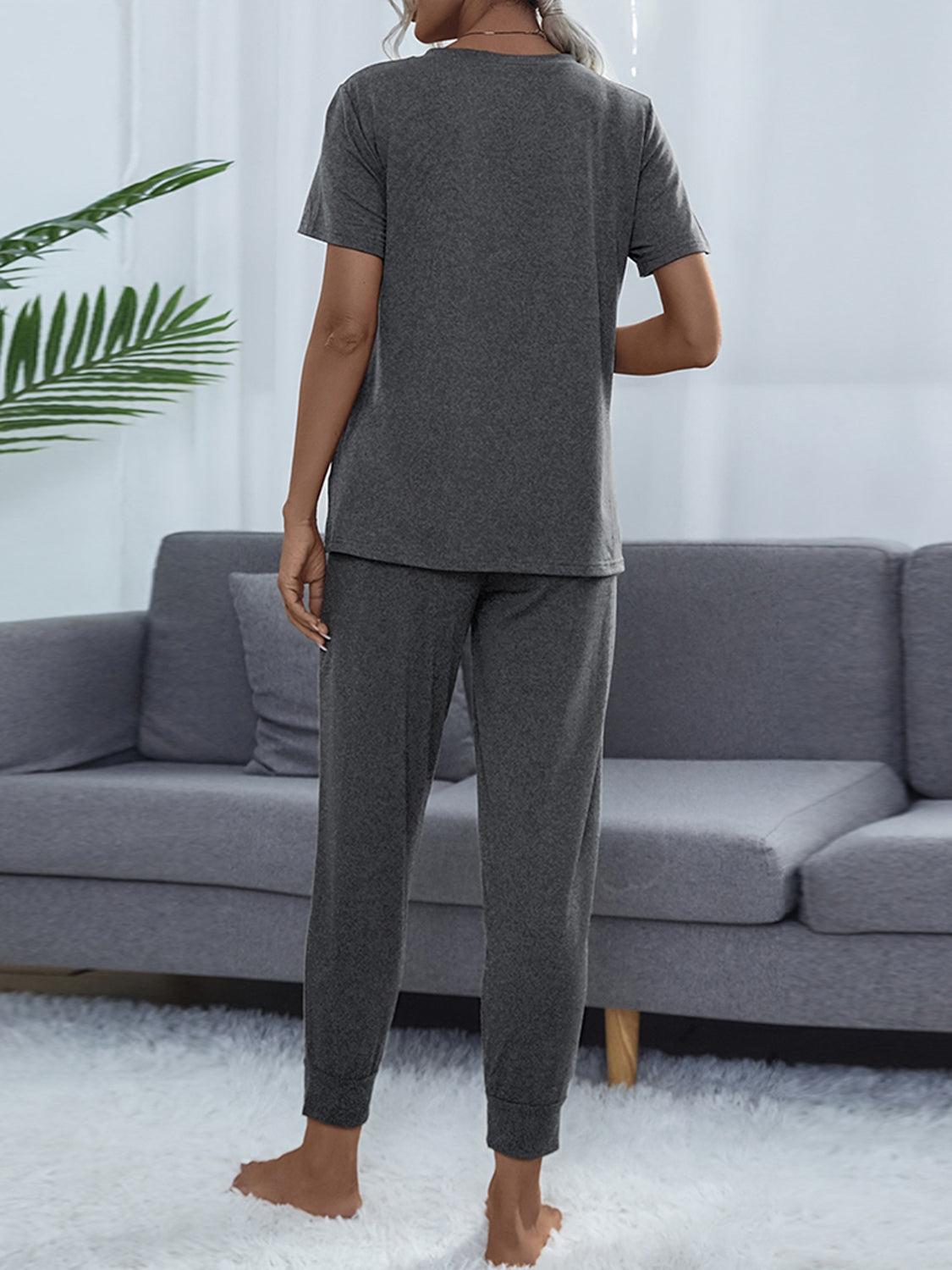 NEWYORKLISTED Intimate Apparel: Person wearing a gray pajama set standing in a living room with a gray sofa and white walls.