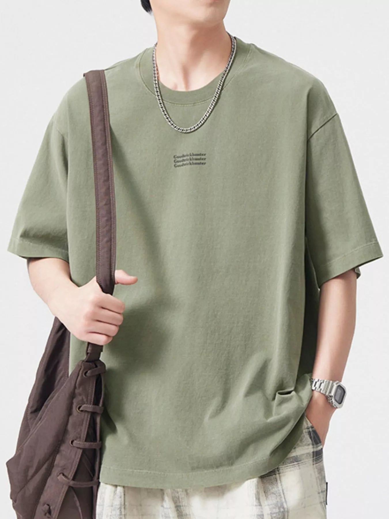 NEWYORKLISTED ATLAS MEN's COLLECTION:  Person wearing a green t-shirt with a subtle logo, holding a brown bag on a plain background