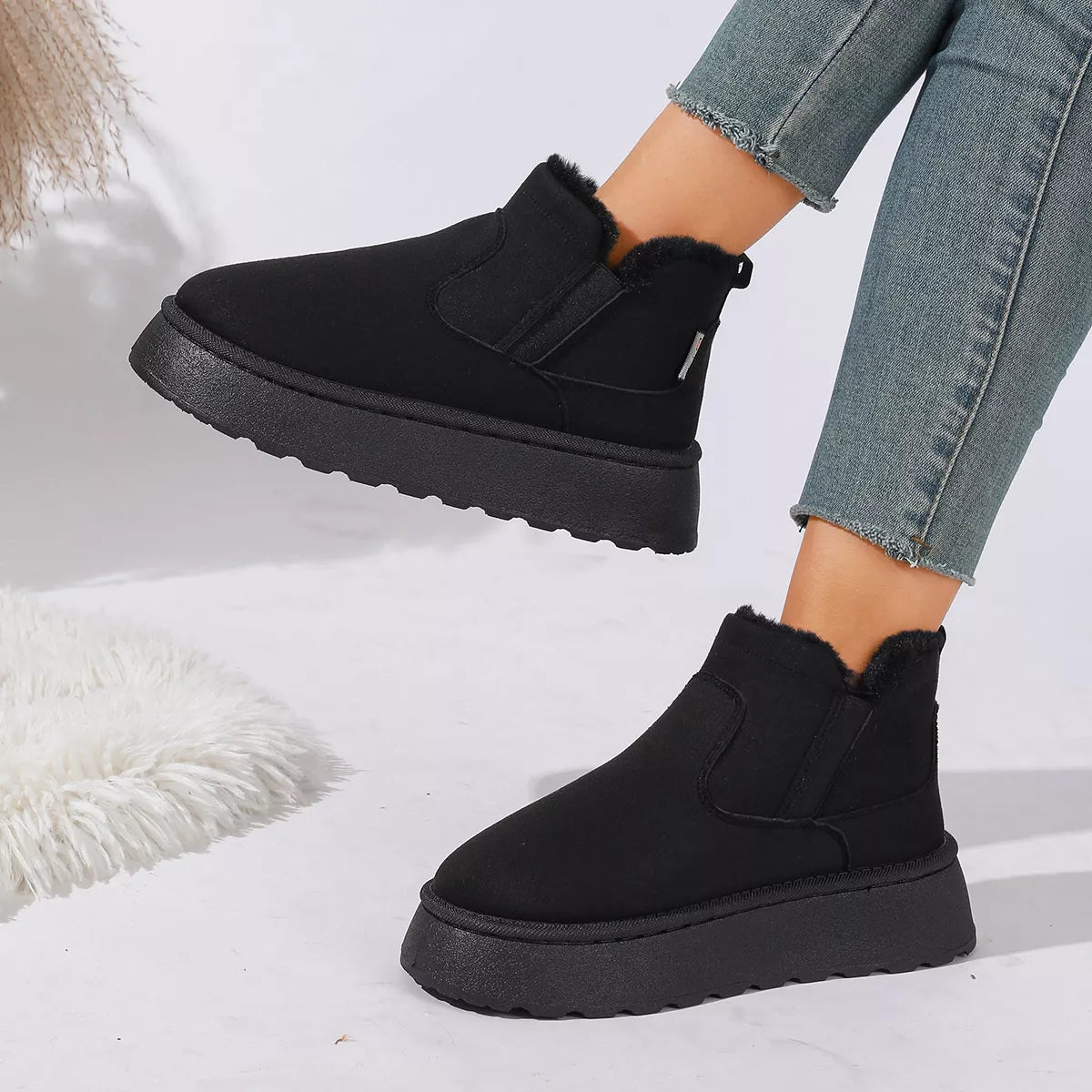 NEWYORKLISTED SHOE COLLECTION: A person wearing black ankle-height slip-on boots with fuzzy trim and chunky black platform soles, paired with light blue frayed-hem jeans, against a clean white studio background.