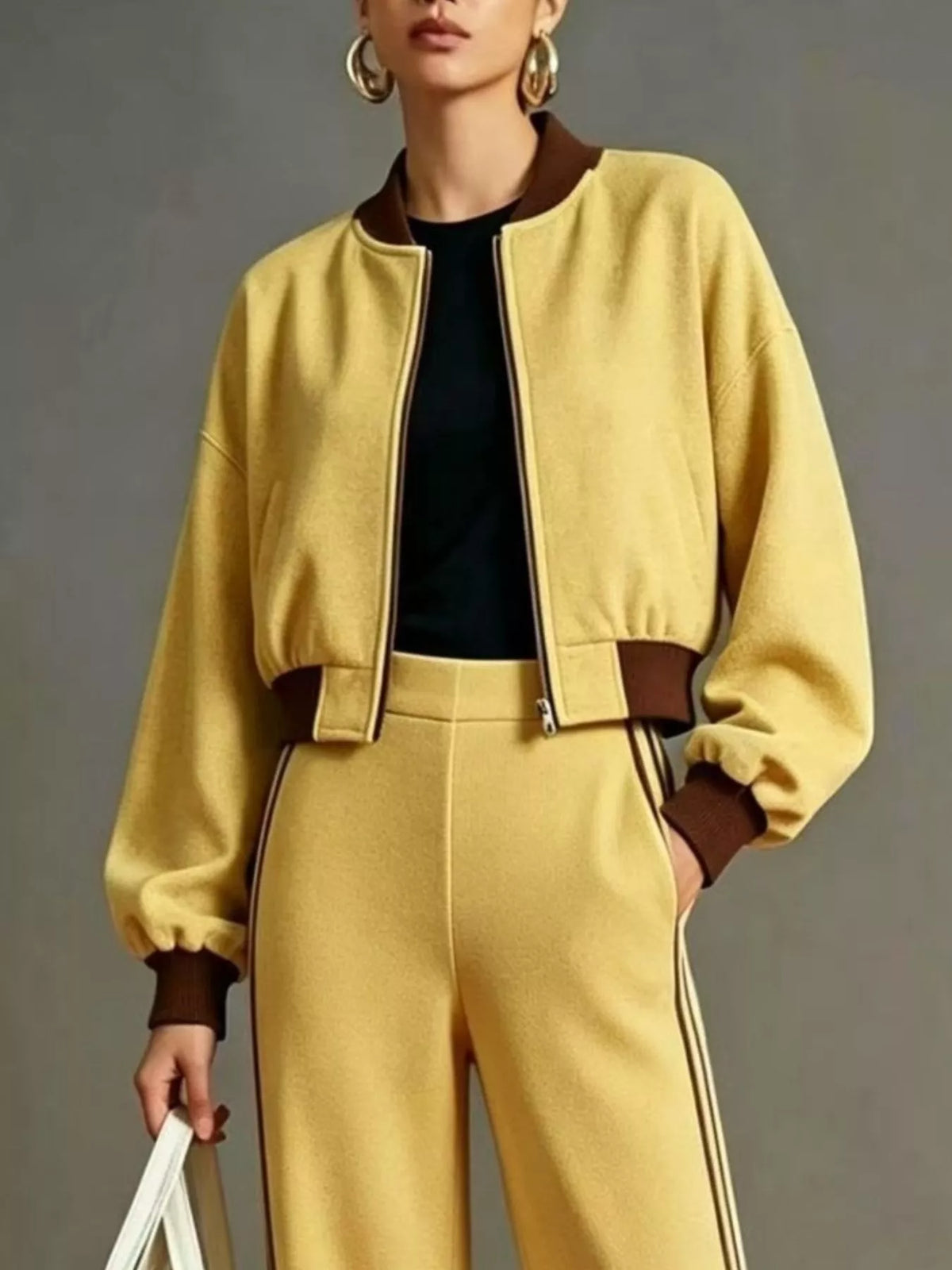 NEWYORKLISTED STYLE COLLECTION: A model wears a matching yellow tracksuit featuring a cropped zip-up bomber jacket with brown ribbed collar, cuffs, and waist detail, paired with matching yellow pants with brown side stripes and a black top underneath.